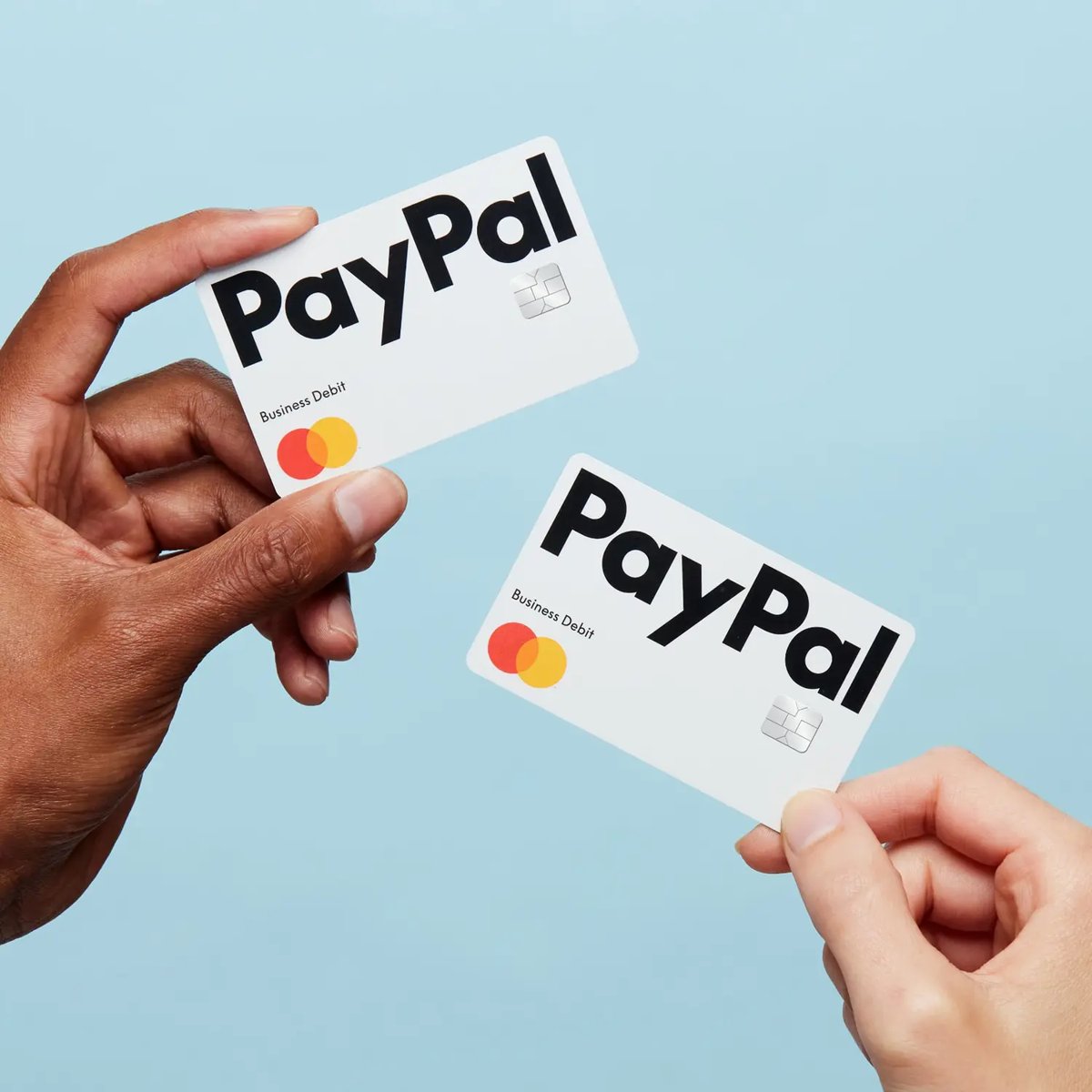 Two white PayPal Business Debit Mastercards held up against a blue background